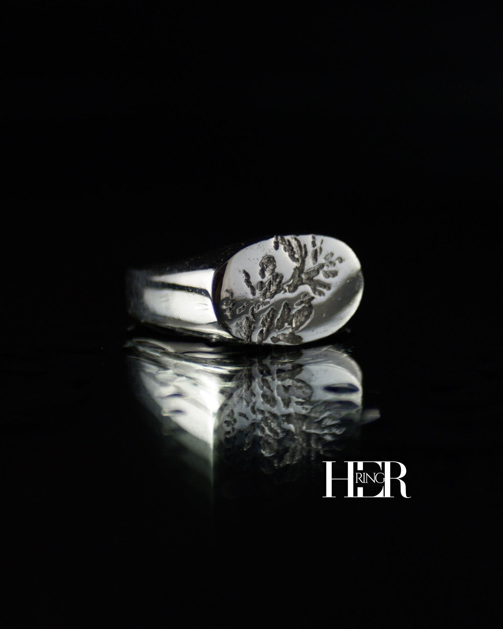 Srebrny sygnet damski, męski o podłużnym kształcie z widocznym odciskiem wrzosu, pokazany na czarnym tle.