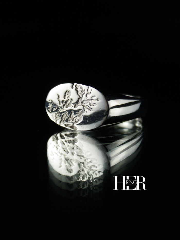 Srebrny sygnet damski, męski o podłużnym kształcie z widocznym odciskiem wrzosu, pokazany na czarnym tle.